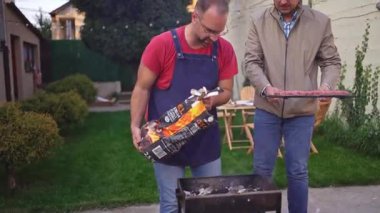 İki arkadaş, biri ızgaraya kömür dökerken diğeri de bir tepsi marine edilmiş et tutarken nefis bir açık hava barbeküsü için hazırlanıyor..