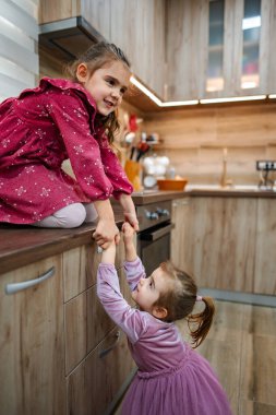 Sıcak ve davetkar bir mutfakta, iki genç kız kardeş hoş bir oyun oynuyorlar. Canlı bir elbise giymiş bir kız kardeş tezgahın üzerine tünemiş, genç olan hevesli bir şekilde ona yardım etmek için uzanır..