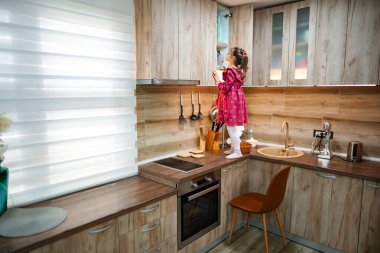 Ahşap dolaplı sıcacık bir mutfakta, pembe elbiseli küçük bir kız tezgahın üzerinde duruyor, merakla uzanıyor. Güneş ışığı içeri akıyor, neşeli atmosfere sıcaklık katıyor..