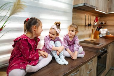 Üç genç kız kardeş mutfak tezgahında oturmuş şakacı sohbetler ediyorlar. Çekici giysiler içinde, sıcacık, aydınlık alanlarında neşe saçıyorlar. İçleri sıcaklık ve kahkahalarla dolu..