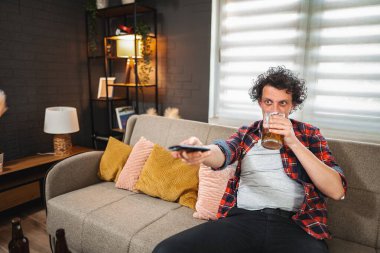 Genç yetişkin bir adam televizyon izlerken bira içiyor, oturma odasında kanepede oturuyor.