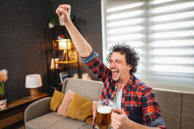 Genç bir adam televizyonda maç izlerken coşkuyla tezahürat yapıyor, elinde bira tutuyor.