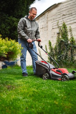 Canlı yeşil çimlerini elektrikli çim biçme makinesiyle biçen bir adam güneşli bir hafta sonunun tadını çıkarıyor. Bahçe düzgünce budanmış çalılar ve yemyeşil çimenler içeriyor, açık hava bakımına olan bağlılığı yansıtıyor..