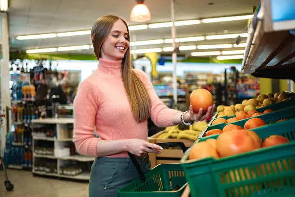 Uzun saçlı, rahat pembe bir kazak giymiş neşeli bir kadın ışıl ışıl ürünlerle dolu bir markette alışveriş yaparken parlak bir elmayı dikkatle inceliyor...