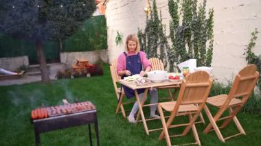 Yeşilliklerle dolu büyüleyici bir arka bahçede, bir kadın neşe içinde barbekü için lezzetli yemekler hazırlıyor. Sıcak ortam ve aile dostu atmosfer eğlenceli bir toplantı için zemin hazırladı..