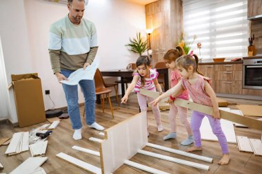 Bir baba ve üç kızı, davetkar yaşam alanlarında yeni bir mobilya inşa etmek için sevinçle iş birliği yapıyorlar. Oda modern, ahşap aksanlı ve doğal ışıkla dolu..