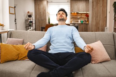 Kahverengi saçlı adam, ferah bir oturma odasındaki kanepede rahatça oturmuş meditasyon yapıyor..