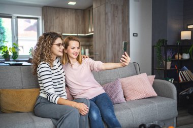 İki kadın bir kanepede oturuyorlar, birlikte selfie çekmek için kol mesafesinde bir akıllı telefon tutuyorlar. Ekrandaki anı yakalarken gülümsüyorlar..