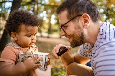 Neşeli bir baba, sonbahar renkleriyle dolu canlı bir parkta meraklı çocuğuyla ılık bir içkiyi paylaşırken gülümser. Ayak altında çıtırdıyor, rahat bir atmosfer yaratıyor..