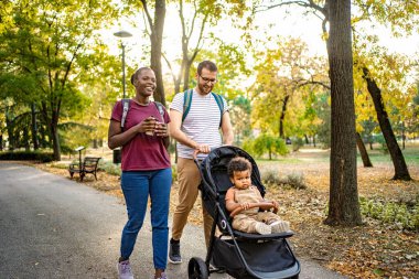 Neşeli bir çift, güneşli bir sonbahar gününün keyfini çıkarırken meraklı bir çocukla bebek arabasını iterken resim parkında el ele yürür. Yapraklar canlı bir zemin oluşturur.