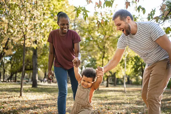 Neşeli bir aile parkta güneşli bir günün tadını çıkarırken, bir çocuk her iki ebeveyn tarafından kaldırılırken neşeyle kıkırdıyor. Yeşil ağaçlar neşeli bağları için canlı bir zemin oluşturuyor..
