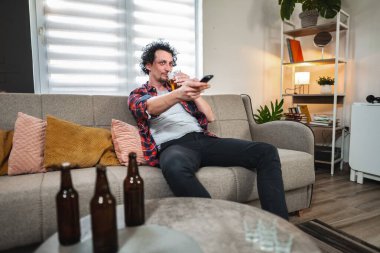 Genç adam bira içiyor ve televizyon seyrediyor, oturma odasında bir kanepede oturuyor.