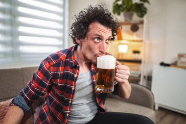 Kıvırcık saçlı genç bir adam evdeki kanepede otururken bir bardak biranın tadını çıkarıyor.