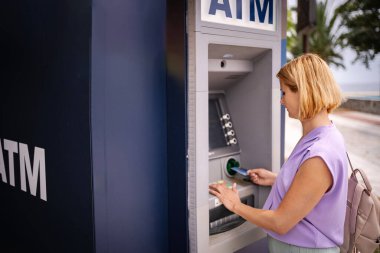 Kadın, güneşli bir Yunan kıyı şeridi boyunca ATM kullanıyor.