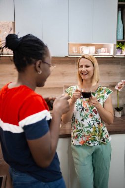 İki kadın, modern bir mutfakta keyifli bir anı paylaşıyor. Her biri elinde bir fincan kahve ve gülümsüyor. Renkli dekor ve güneş ışığı dostluk ve kahkaha için davetkar bir atmosfer yaratır..