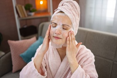 Bornoz ve havlu giyen genç bir kadın evde yüzüne kozmetik maske takıyor.