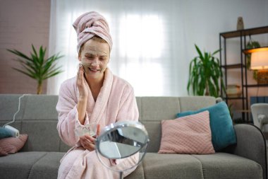 Gülümseyen genç bir kadın yüzüne kilden bir maske takıyor aynaya bakıyor, evdeki kanepede oturuyor.