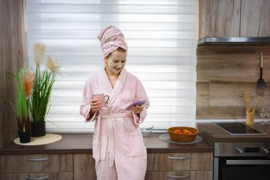 Bornoz ve yüz maskesi takan, akıllı telefon kullanan ve mutfakta kahve içen genç bir kadın.