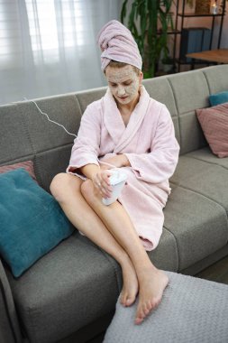 Evdeki kanepede oturan kadın, bornoz ve yüz maskesi takıyor, saç aldırmak için IPL cihazı kullanıyor.