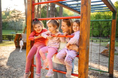 Gülümseyen ve mutlu anne, anaokulu kızı ve ikizleriyle oyun bahçesinde..