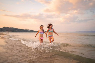 İki neşeli kız kardeş güneş batarken su sıçratarak kıyı şeridi boyunca koşuyor..