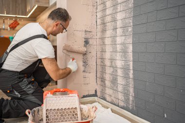 Bir adam beyaz boyalı tuğla bir duvar boyuyor. Boyayı yüzeye eşit olarak uygulamak için bir silindir kullanıyor, kırmızı tuğlaları tamamen kaplıyor. Duvar orijinal kırmızı renginden taze, temiz beyaz rengine dönüşüyor..