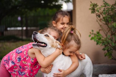 Üç mutlu çocuk açık havada dost canlısı bir köpekle sevgi dolu bir kucaklaşmayı paylaşıyor..