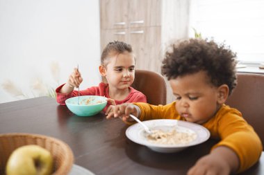 İki çocuk, sıcak bir mutfakta, yemek masasında yemek yiyorlar..