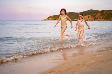 İki kız, sakin bir sahilde dalgaların arasından neşeyle sıçrıyor..