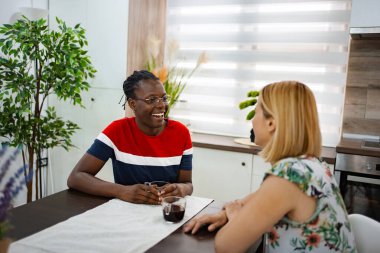 İki arkadaş, parlak bir mutfakta, ahşap bir masada canlı bir sohbete girişiyor. Güneş ışığı pencereden akar ve sıcak ve davetkar bir atmosfer yaratır..