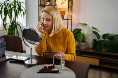 Yeşillik dolu davetkar bir mekanda, bir kadın, nazikçe öğleden sonra ışığında, makyaj ürünleriyle çevrili, ahşap bir masada otururken güzelliğini dikkatlice geliştirir..