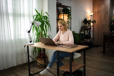 Bir kadın, yeşil ve yumuşak ışıklandırma ile çevrili büyüleyici bir ev ofisinde dizüstü bilgisayarına odaklanır ve bu da rahatlatıcı bir atmosfer yaratır. Huzurlu bir yerde çalışmaktan hoşlanıyor..