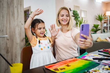 Bir anne ve kızı, renkli sanat eserleri ve mutfak malzemeleriyle çevrili bir video görüşmesi sırasında kameraya mutlu bir şekilde el sallıyor..