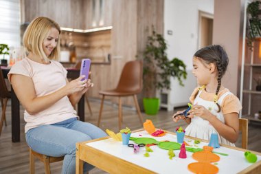 Bir anne, kızı rahat evlerindeki bir masada renkli bir mankenle oynarken telefonuyla bir an yakalar..