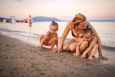 Bir anne gün batımında ikiz kızlarıyla kumda oynar..