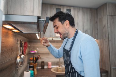 Bir adam mutfakta elinde kaşıkla dikiliyor. Elindeki işe odaklanarak yemek hazırlıyor ya da pişiriyor gibi görünüyor. Mutfak iyi aydınlatılmış ve derli toplu. Arka planda mutfak aletleri görünüyor..