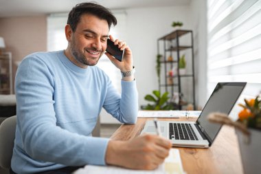 Bir adam bir masada oturuyor, cep telefonuyla sohbet ediyor. Odaklanmış ve özenli görünüyor, muhtemelen iş yapıyor ya da kişisel bir telefon görüşmesi yapıyor..