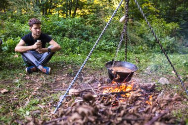 Genç yürüyüşçü ormandaki bir kamp ateşinde yemek pişirirken dinleniyor.