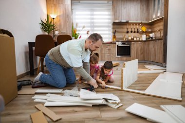 Sıcak ahşap tonlarla dolu modern bir mutfakta bir baba, iki çocuğuyla birlikte çalışarak mobilya montajı yapar..