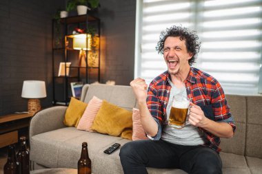 Evde televizyonda spor maçı izlerken bira içip tezahürat yapan genç bir adam.