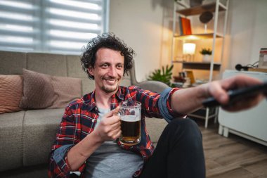 Genç adam televizyon seyrediyor ve bira içerken kanal değiştiriyor, oturma odasında yerde oturuyor.