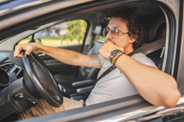 Kıvırcık saçlı, güneş gözlüklü, araba süren ve sigara içen yakışıklı bir adam..