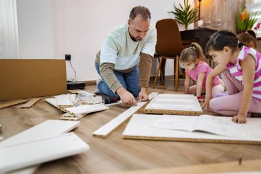 Bir adam iki çocuğun doğal ışıkla dolu parlak bir oturma odasında mobilya monte etmesine yardım ediyor. Kahkaha ve takım çalışması havayı doldurur. Talimatlara dikkatle uyarlar ve sürecin tadını çıkarırlar..