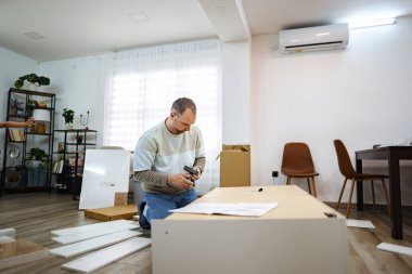 Odaklanmış bir adam yerde diz çöker ve şık bir oturma odasında mobilyaları dikkatlice birleştirir. Güneş ışığı perdelerin arasından süzülüp sıcak ve davetkar bir atmosfer yaratır..