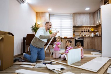 Bir adam, üç küçük kızı hevesle izlerken ve yardım ederken yerde diz çöküp mobilyaları birleştiriyor. Sıcak mutfak ortamı aralarında bir bağ ve yaratıcılık hissi uyandırıyor..