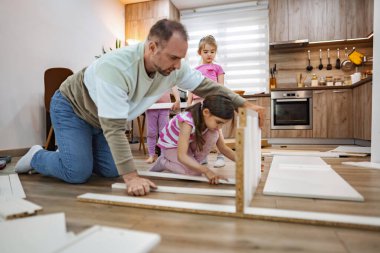 Bir baba ve iki kızı şık mutfaklarında yeni mobilyalar yapmaya odaklanmışlar. Kızlar birlikte güzel vakit geçirirken parçaları birleştirerek yardım ediyorlar..