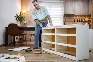 Odaklanmış bir adam, çağdaş bir oturma odasında parlak beyaz bir dolap oluşturmak için gayretle çalışır. Etrafı dağınık alet ve parçalarla çevrili vazifesinde kararlılık ve beceri gösteriyor..