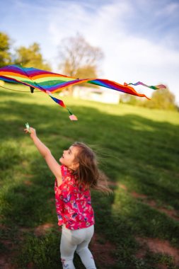 Genç bir kız yemyeşil çimlerin arasında koşuyor, gülüşü gökkuşağı uçurtmasının kuyruğunu tutarken havayı dolduruyor. Güneş parlıyor, dışarıda eğlence için güzel bir gün yaratıyor..