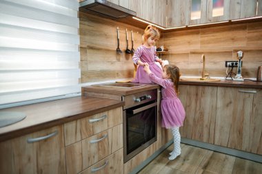 Işıkla dolu rahat bir mutfakta, iki küçük kız çevrelerini keşfediyor. Biri tezgahın üstünde dururken diğeri ona yardım ediyor. İkisi de aynı pembe elbiseleri giyip neşeyle gülüyor..