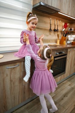 İki genç kız kardeş bir mutfakta keyifli bir an yaşıyor, biri tezgahın üzerine tünemiş, diğeri de onun yükselişine yardım ediyor..